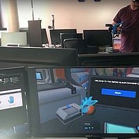 Ein Computerbildschirm mit einer virtuellen Realität, dahinter ein Mensch mit einer VR Brille