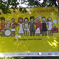 Poster of migrantas with different people, stretched between trees. On it is written: A dignified life for all