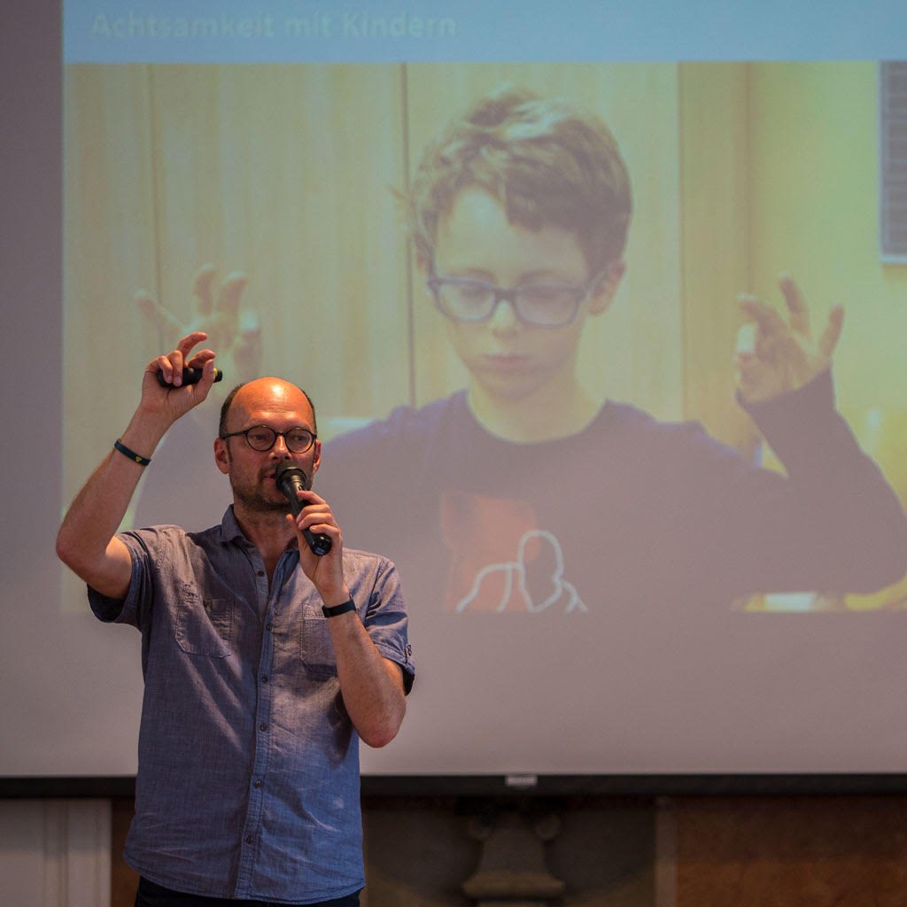 Auf dem Foto ist Nils Altner bei einem Vortrag zu sehen. Im Hintergrund ist eine Leinwand zu sehen mit dem Foto von einem Kind darauf, das die Augen geschlossen hat und mit seinen Fingern bestimmte Übungen macht.  