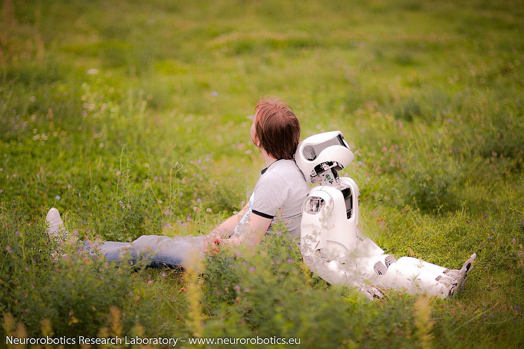 Ein Mann und ein weißer Roboter sitzen mit dem Rücken aneinandergelehnt auf einer Wiese