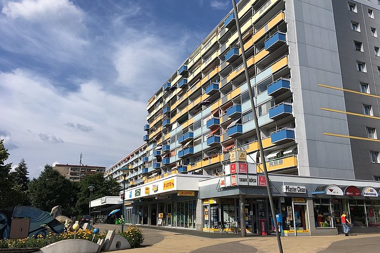 The picture shows an apartment block and the forecourt, the lowest floor of the building has stores.