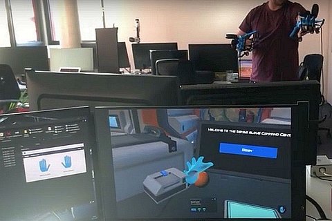 Ein Computerbildschirm mit einer virtuellen Realität, dahinter ein Mensch mit einer VR Brille