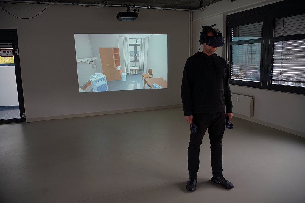 Ein Mann mit VR-Brille in einem Raum, im Hintergrund auf einem Beamerbild ein virtuelles Patientenzimmer