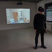 Ein Mann mit VR-Brille in einem Raum, im Hintergrund auf einem Beamerbild ein virtuelles Patientenzimmer