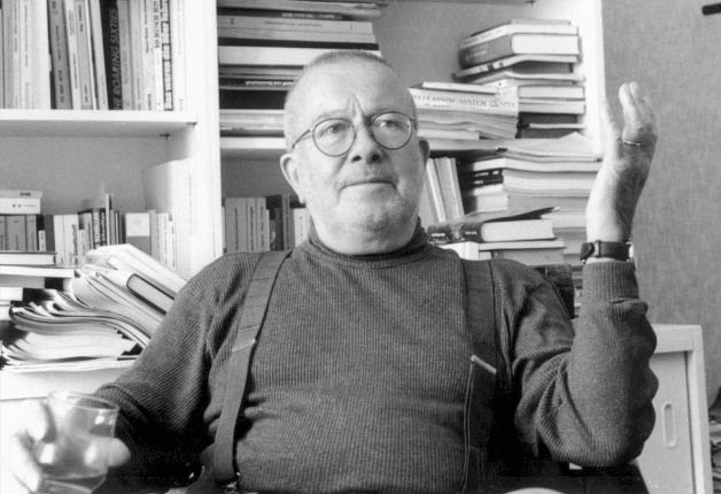 C.W. Müller mit Hosenträgern vor einem Bücherregal sitzend