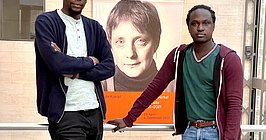 Zwei Männer an eine Balustrade gelehnt vor einem Porträt von der jungen Angela Merkel