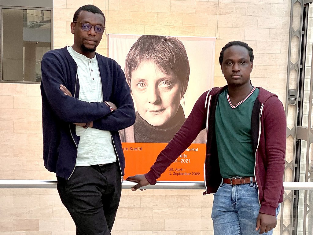 Zwei Männer an eine Balustrade gelehnt vor einem Porträt von der jungen Angela Merkel