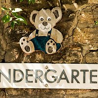 Auf einer Mauer ein Schild mit der Aufschrift Kindergarten und darüber ein Teddy aus Holz