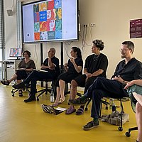 Die Podiumsgäste sitzen nebeneinander in einem Seminarraum der ASH Berlin. Im Hintergrund befindet sich ein großer Bildschirm auf dem das Cover des Buches "Wie geht es dir?" zu sehen ist.