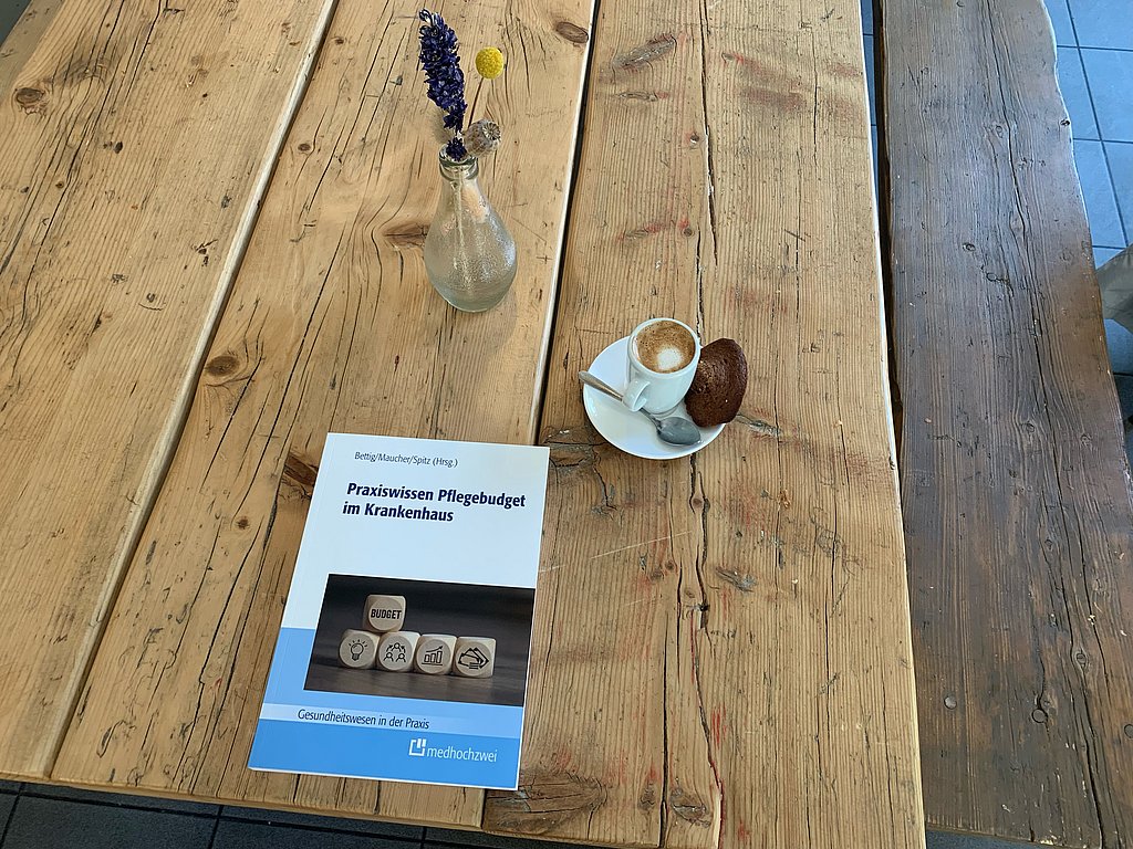 Das Buch auf einem Holztisch mit einem Kaffee und einem Blumenstrauß daneben