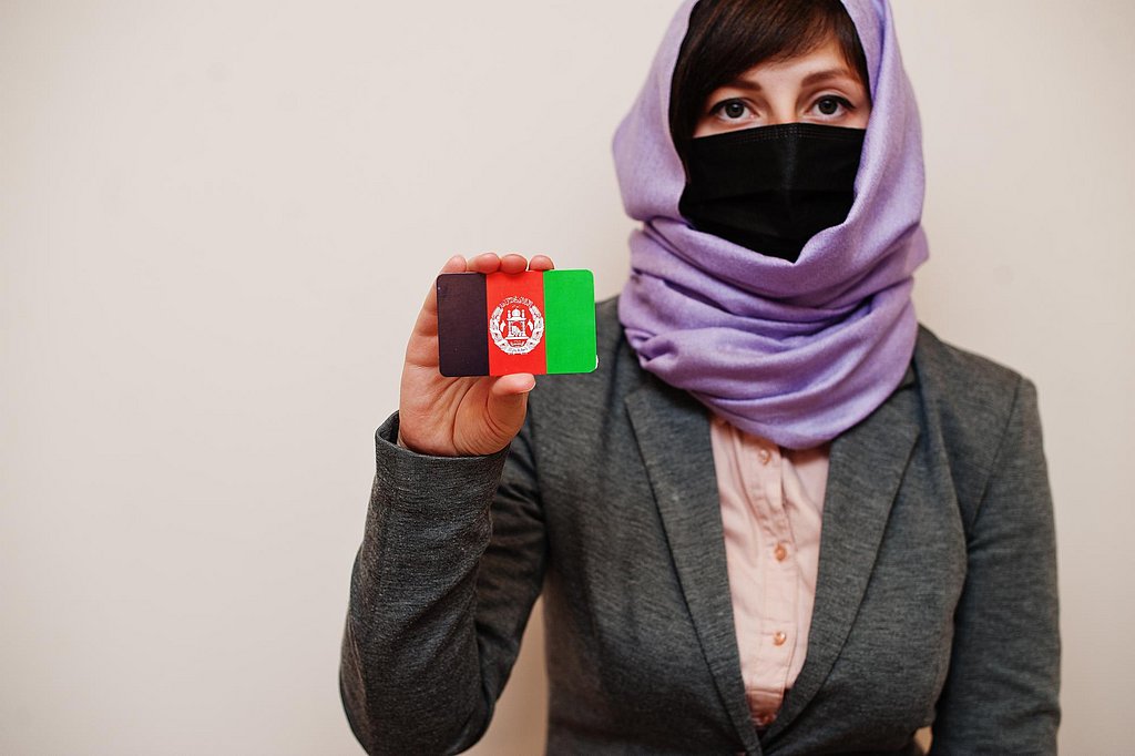 Eine Frau mit lila Kopftuch und schwarzem Mund- Nasenschutz hält eine Karte mit der afghanischen Flagge hoch