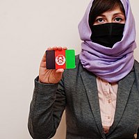 Eine Frau mit lila Kopftuch und schwarzem Mund- Nasenschutz hält eine Karte mit der afghanischen Flagge hoch