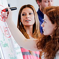 Eine Studentin schreibt mit rotem Marker auf ein Flip-Chart Papier. Eine Kommilitonin steht hinter ihr und hat den Blick auf sie gerichtet.