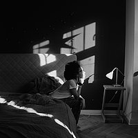Schwarz-Weiß Fotografie einer jungen Frau, die auf einem Bett sitzt. Ihr scheint sie Sonne ins Gesicht. 