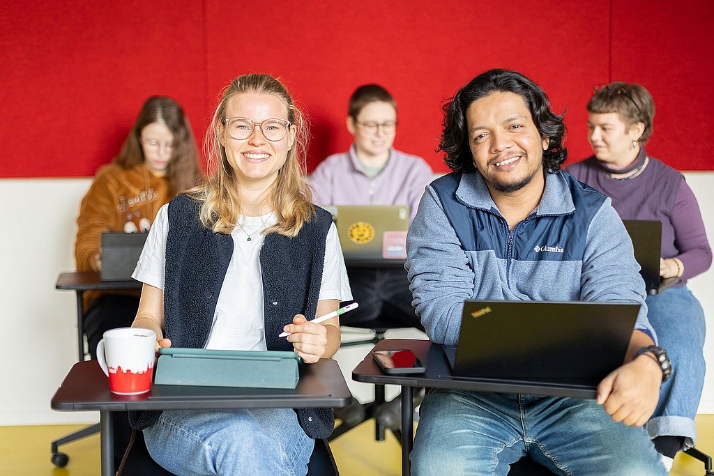 Studierende der ASH Berlin sitzen an Tischen in einem Seminarraum der Hochschule und lächeln in die Kamera