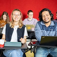 Studierende der ASH Berlin sitzen an Tischen in einem Seminarraum der Hochschule und lächeln in die Kamera