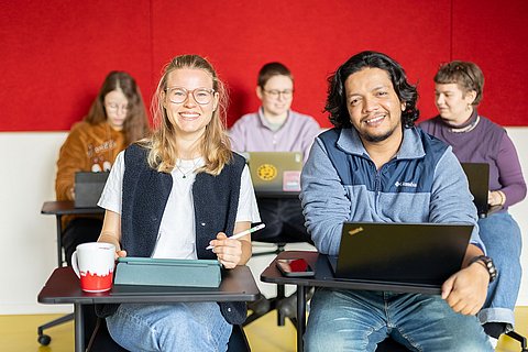 Studierende der ASH Berlin sitzen an Tischen in einem Seminarraum der Hochschule und lächeln in die Kamera