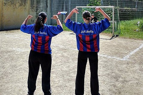 Das Bild zeigt zwei Fußballspielerinnen in JAA Trikots von hinten, die auf ein Tor blicken, in dem ein junger Torwart auf einen Schuss wartet.