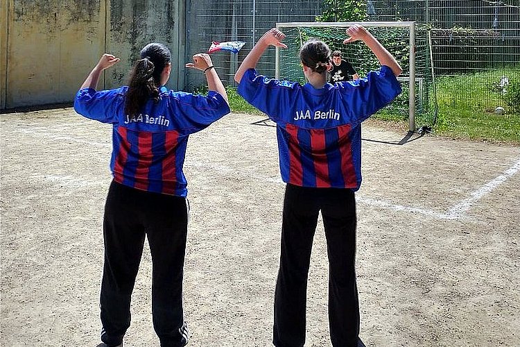 Das Bild zeigt zwei Fußballspielerinnen in JAA Trikots von hinten, die auf ein Tor blicken, in dem ein junger Torwart auf einen Schuss wartet.