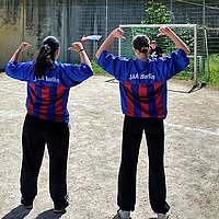 The picture shows two female soccer players in JAA jerseys from behind, looking towards a goal where a young goalkeeper is waiting for a shot.