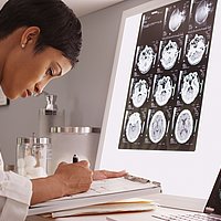 Eine Frau im weißen Kittel und dunkler Hautfarbe arbeitet am Schreibtisch, vor ihr hängen Röntgenaufnahmen