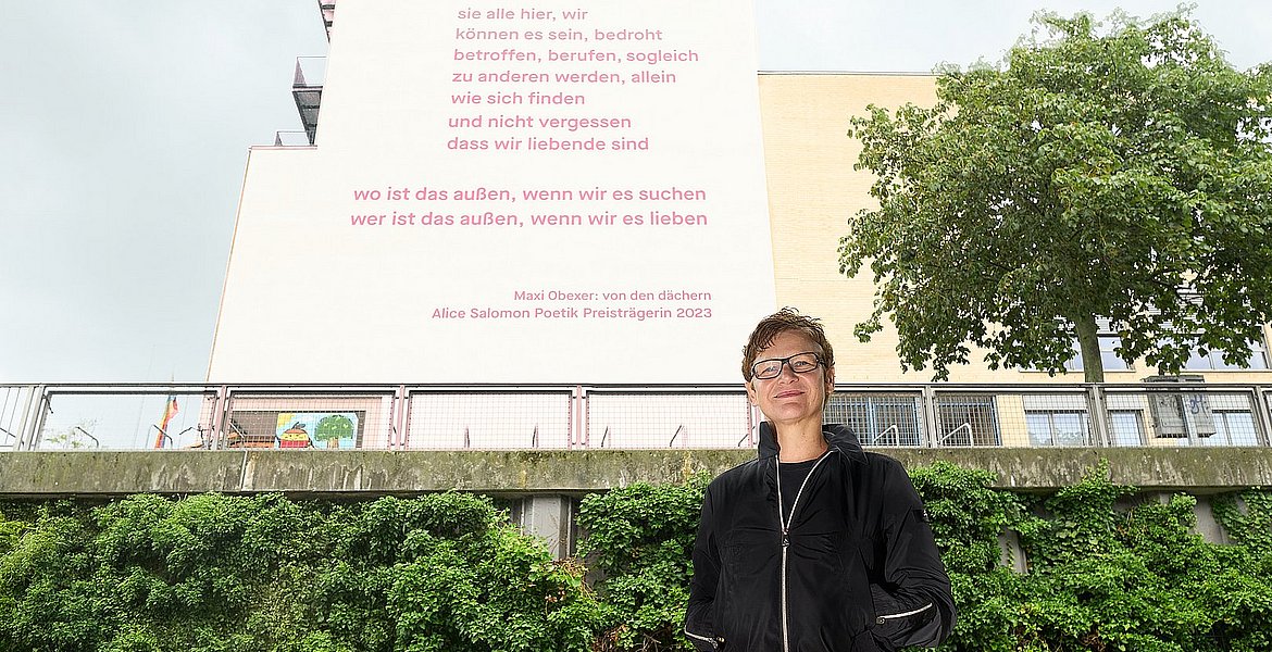 The photo shows Maxi Obexer in front of the south façade of ASH Berlin, on which Obexer's poem "von den dächern" can be read.