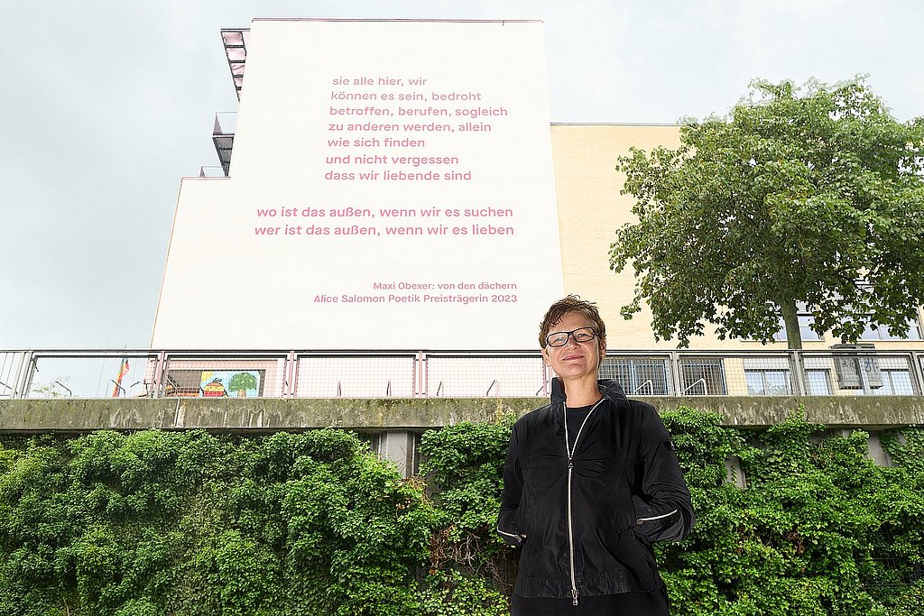 The photo shows Maxi Obexer in front of the south façade of ASH Berlin, on which Obexer's poem "von den dächern" can be read.