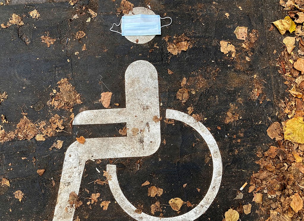 Ein Rollstuhl auf Asphalt gemalt für einen Behindertenparkplatz. Auf dem Kopf liegt ein Mundschutz