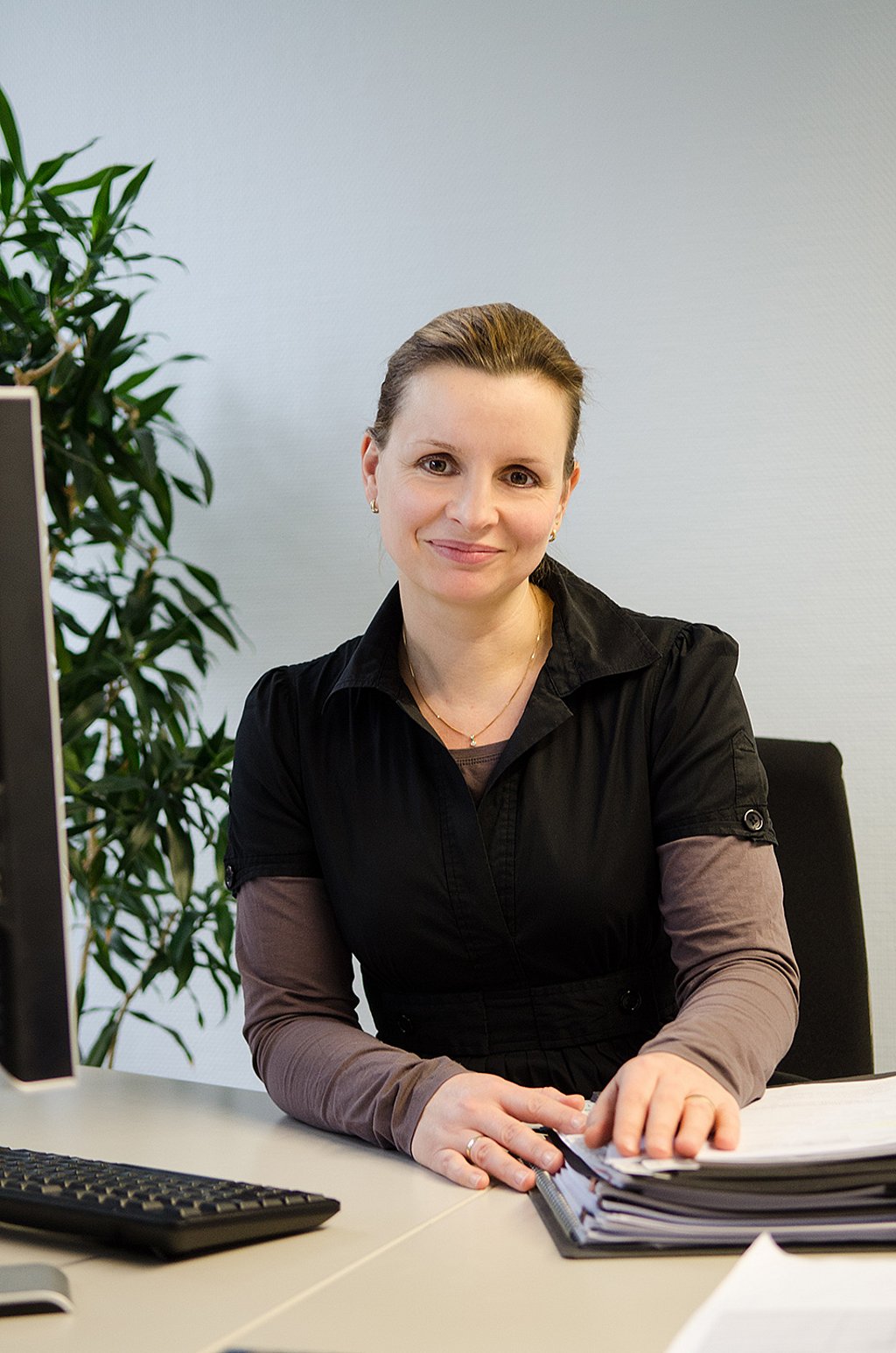 Katja Thielemann sitzt an ihrem Computer am Schreibtisch, hat die Hände auf eine Aktenmappe gelegt und ihren Blick nach vorne gerichtet.