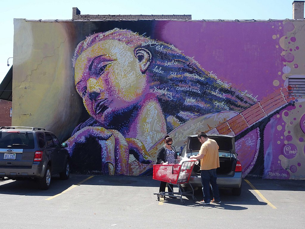Zwei Personen räumen auf einem Parkplatz ihre Einkäufe in ihr Auto. Dahinter ist an der Hausfassade riesig groß das Portrait einer Geigerin gemalt.
