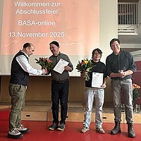 Im Audimax der ASH Berlin stehen vier Personen nebeneinander. Sie schauen in die Kamera und halten Blumen in der Hand. 