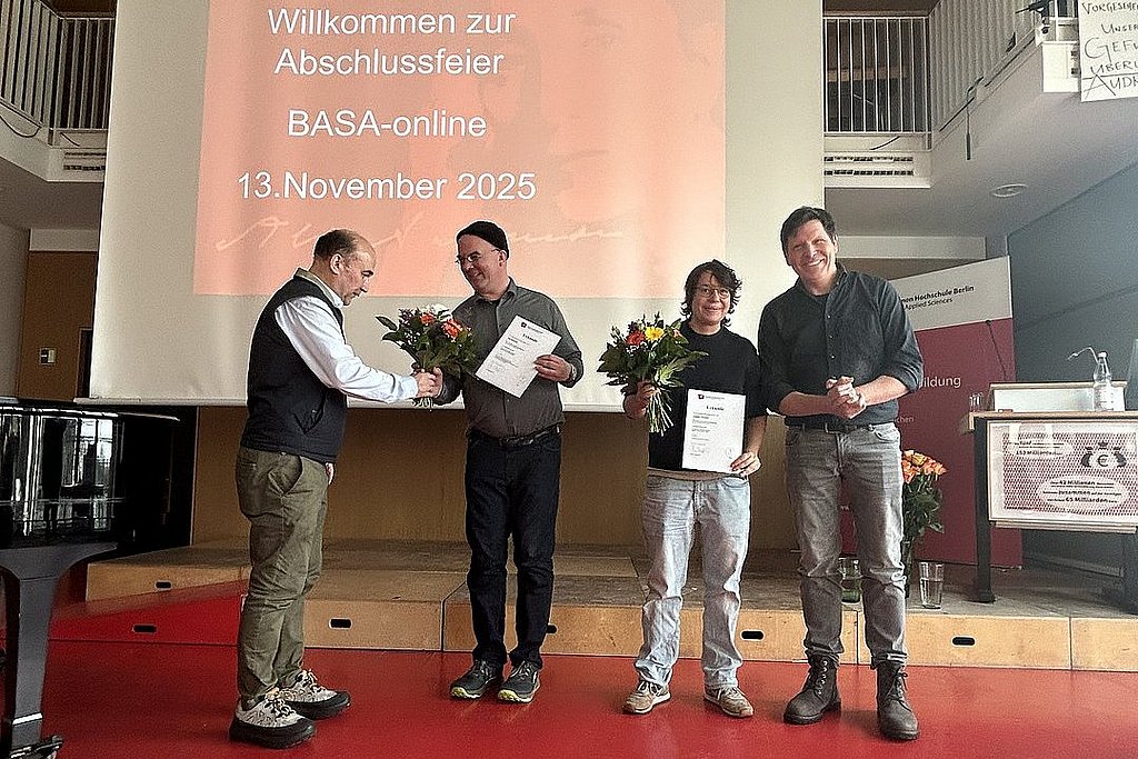 Im Audimax der ASH Berlin stehen vier Personen nebeneinander. Sie schauen in die Kamera und halten Blumen in der Hand. 