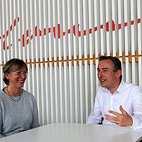Gudrun Piechotta-Henze und Lutz Schumacher sitzen an einem Tisch in der Mensa der ASH Berlin vor einer Holzwand mit der Unterschrift von Alice Salomon
