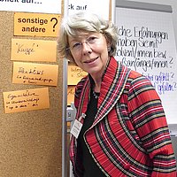 Portraitaufnahme von Ingrid Stahmer, die vor einer orangenen und grünen Kartons bestückten Pinnwand steht.