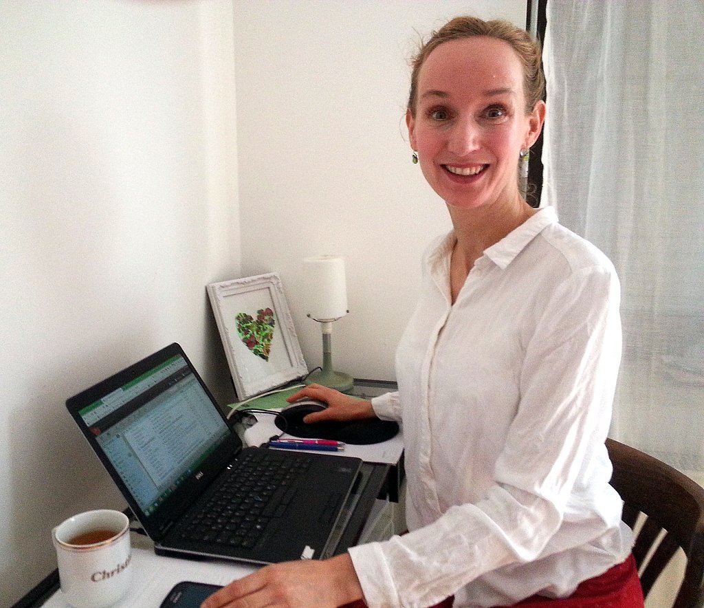 Christine Blümke sitzt an einem kleinen Schreibtisch mit Laptop in ihrer Wohnung vor einer weißen Wand