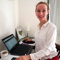 Christine Blümke sitzt an einem kleinen Schreibtisch mit Laptop in ihrer Wohnung vor einer weißen Wand