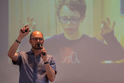 Auf dem Foto ist Nils Altner bei einem Vortrag zu sehen. Im Hintergrund ist eine Leinwand zu sehen mit dem Foto von einem Kind darauf, das die Augen geschlossen hat und mit seinen Fingern bestimmte Übungen macht.  