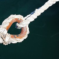 Ein verschlissenes Seil mit einem rostigen Ring hängt über dem Wasser, von oben gehalten von einem dünneren, intaktem Seil.