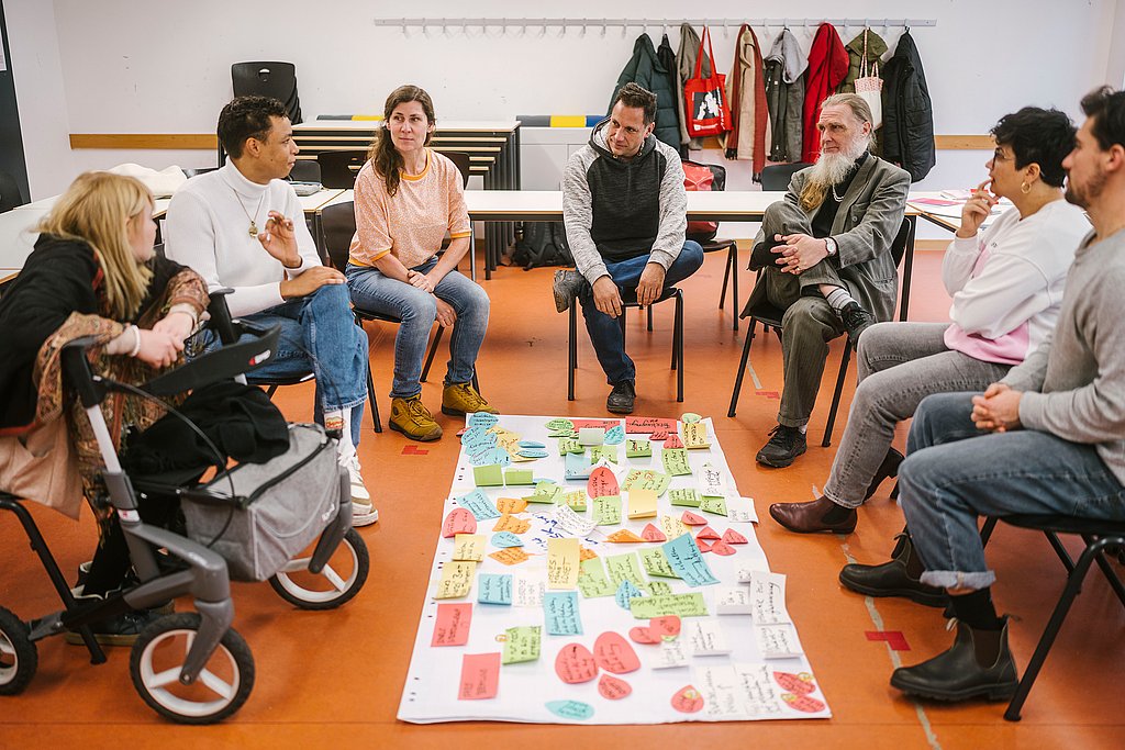 Menschen sitzen in einem Seminarraum im Kreis auf Stühlen und einem Rollstuhl, in der Mitte auf dem Fußboden ein Flipchart-Blatt mit bunten beschriebenen Moderationskarten