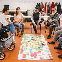 Menschen sitzen in einem Seminarraum im Kreis auf Stühlen und einem Rollstuhl, in der Mitte auf dem Fußboden ein Flipchart-Blatt mit bunten beschriebenen Moderationskarten