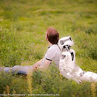 Ein Mann und ein weißer Roboter sitzen mit dem Rücken aneinandergelehnt auf einer Wiese