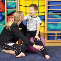 Eine Physiotherapeutin hat ihre Hand platziert auf dem Unterschenkel eines kleinen Jungen, der auf eine Medizinball sitzt. Sie zeigt ihm Übungen.