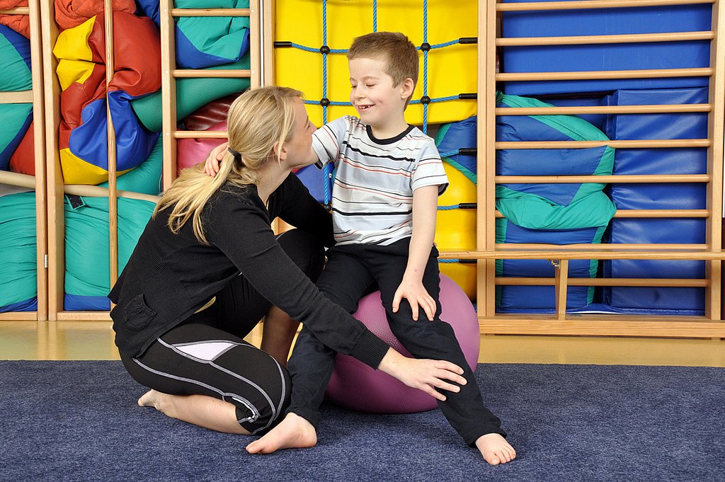 Eine Physiotherapeutin hat ihre Hand platziert auf dem Unterschenkel eines kleinen Jungen, der auf eine Medizinball sitzt. Sie zeigt ihm Übungen.