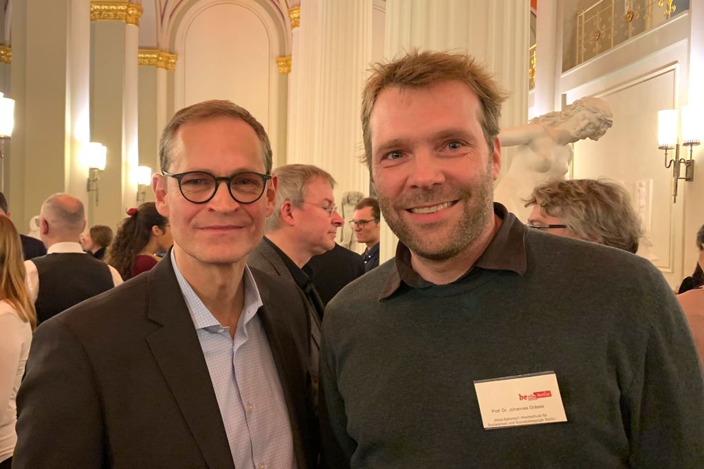 Vergrößern: Die beiden Personen blicken in die Kamera, im Hintegrund Säulen