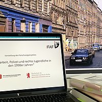 Ein Laptop auf dessen Bildschirm der Projektname steht, rechts daneben ein Bücherstapel