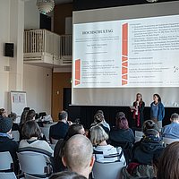 Das Foto zeigt den Audimax. Gesine Bär und Bettina Völter stehen vor der Leinwand mit dem Programm des Hochschultags.