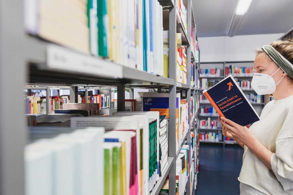 Vergrößern: Eine Frau steht mit Mundschutz vor einem Bücherregal und liest in einem Buch