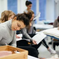 Eine Studentin schreibt etwas auf einem Tisch vor ihr, im Hintergrund liegt eine PErson auf einer Liege