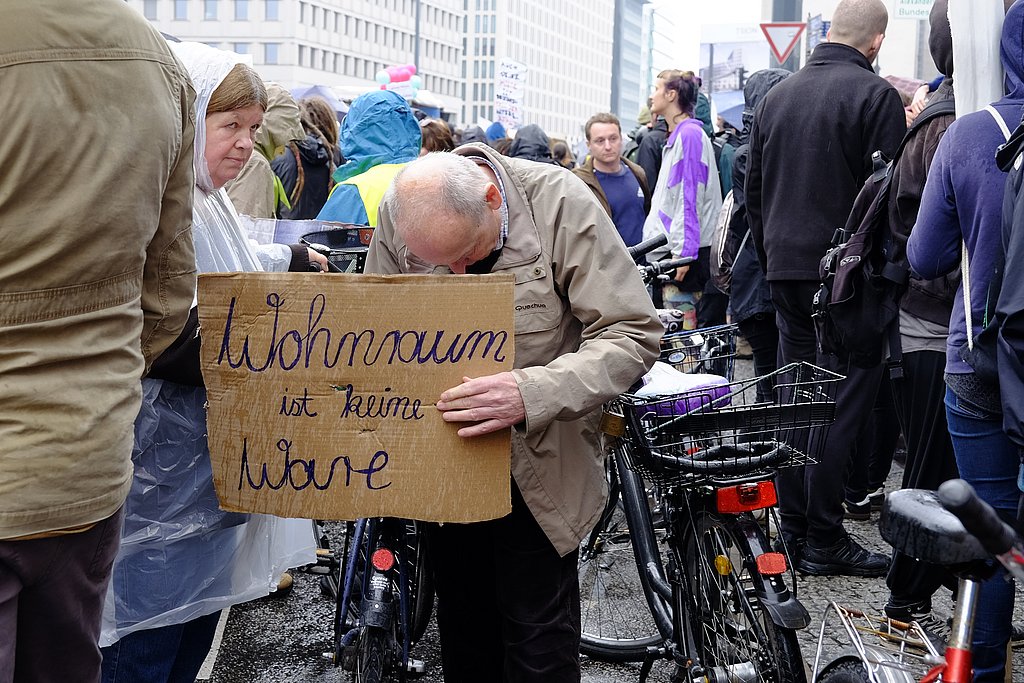 Ein alter Mann mit schütterem Haar und gebeugtem Kopf hält ein Pappschild in der Hand mit der Aufschrift: Wohnraum ist keine Ware