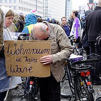 Ein alter Mann mit schütterem Haar und gebeugtem Kopf hält ein Pappschild in der Hand mit der Aufschrift: Wohnraum ist keine Ware